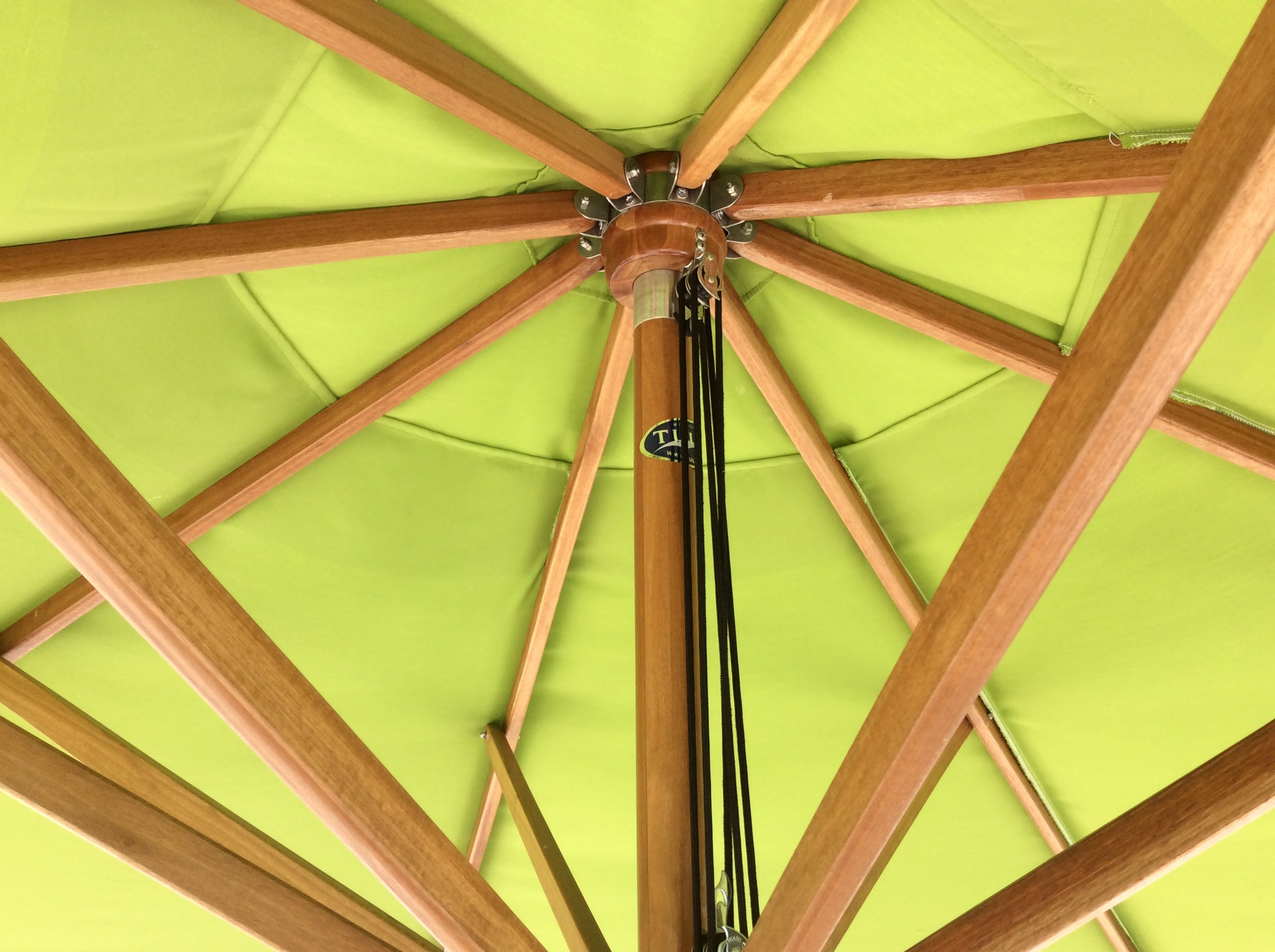 Close-up of a wooden color patio umbrella with a green canopy. Image is showing the details of the inside of the umbrella up-close. There is a pulley system with a black cord and eight arms to the umbrella. Canopy is a bright green chartreuse color.